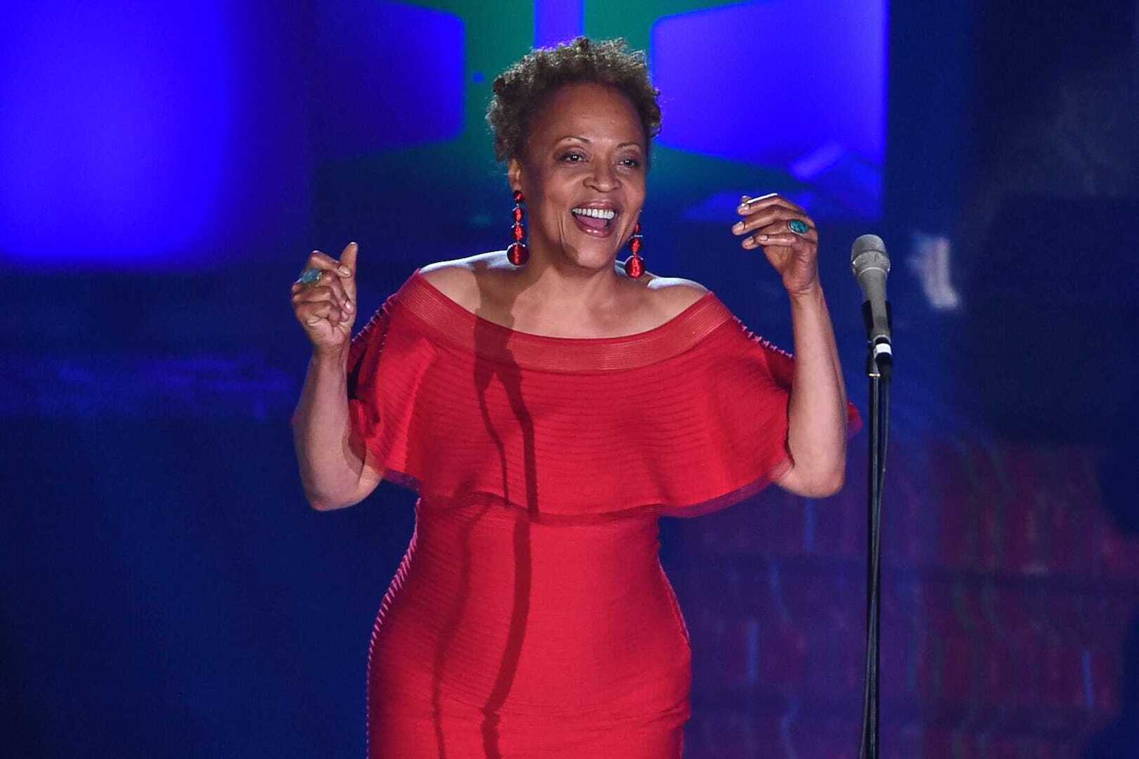 Cassandra Wilson beams joyfully in a red off-shoulder dress on stage, arms raised near a microphone.